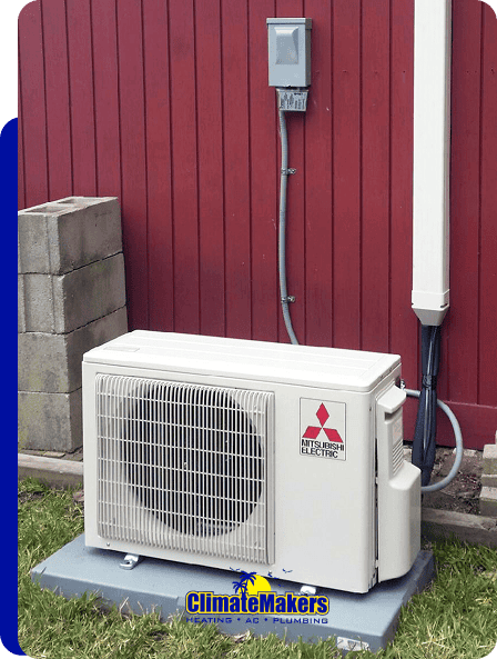 ClimateMakers outdoor Mitsubishi electric HVAC unit sits on a concrete pad by a red wooden wall, with electrical wiring connected above. The ClimateMakers logo is clearly visible in the image.