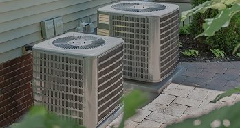 Two outdoor climatemakers air conditioning units are positioned side by side on a paved area next to a house.