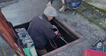 Two climatemakers work on pipes and equipment in an open utility hole on a concrete surface, with tools and a component nearby.